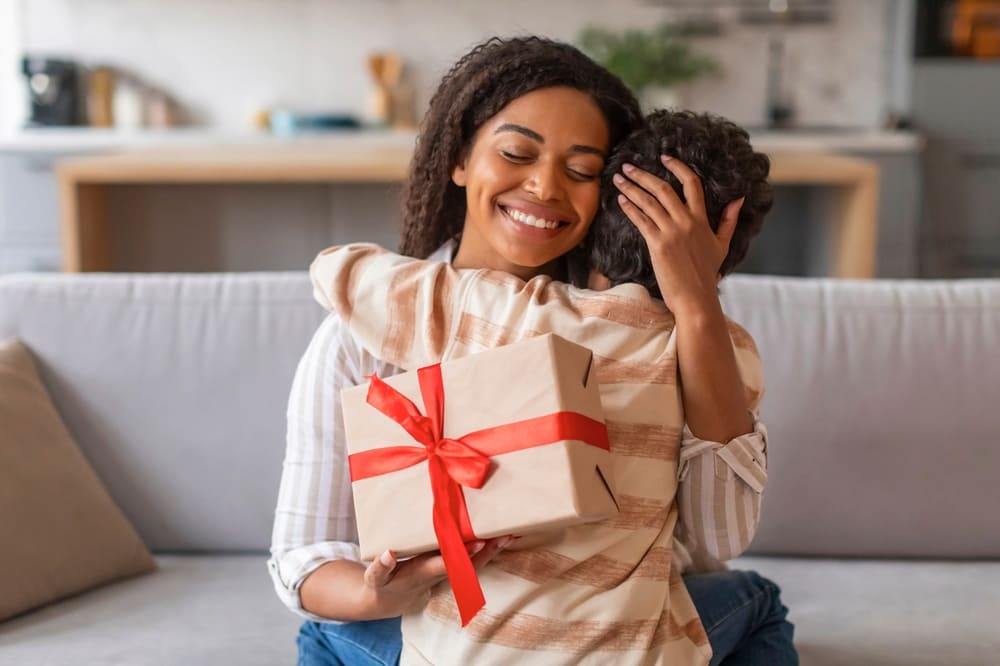 Dia das mães Dia das mães, uma mulher de pele negra, cabelos castanhos, olhos castanhos, jovem, sorrindo ao abraçar seu filho ao granhar um presente de dia das mães.